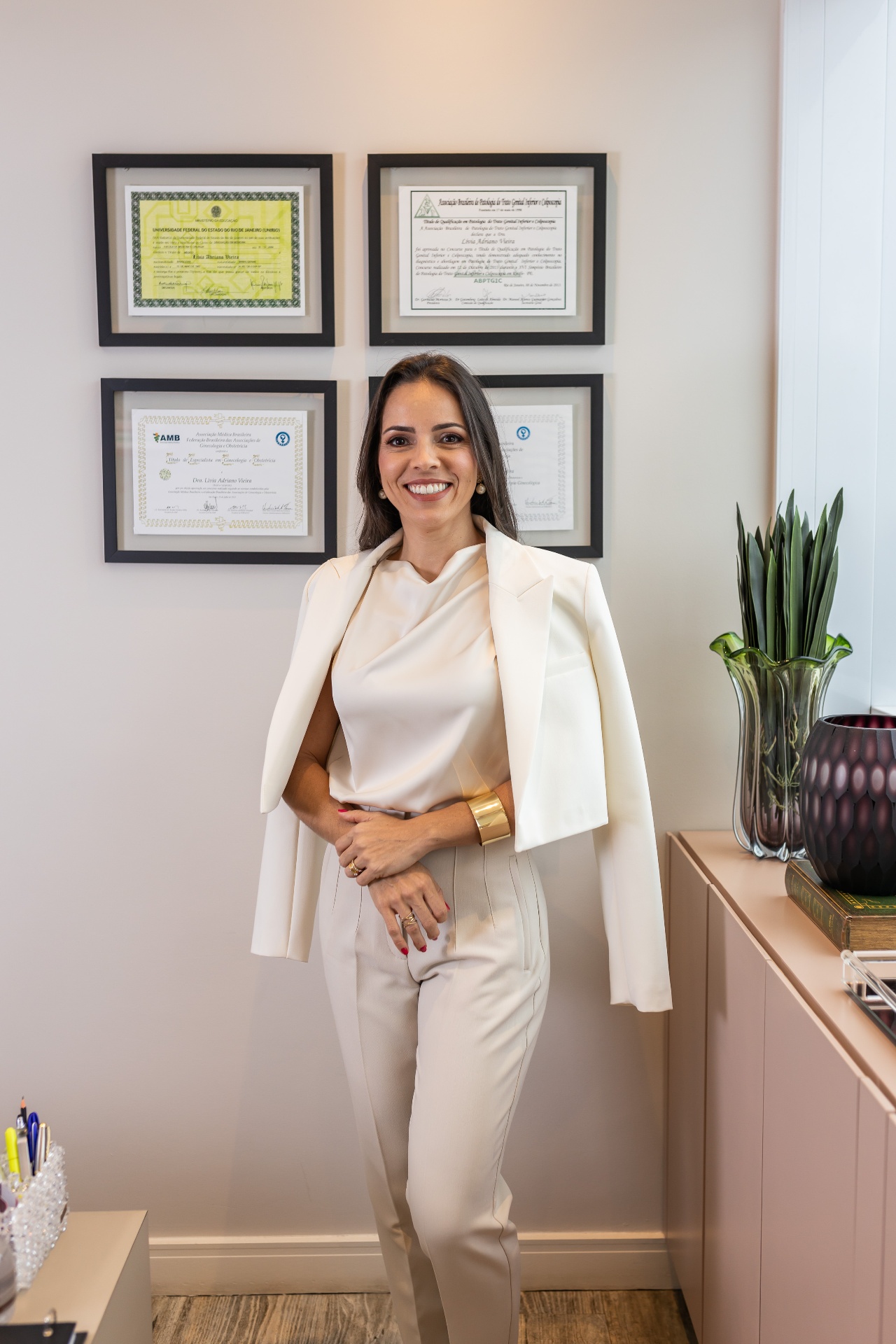 Dra. Lívia Vieira em seu consultório, ao lado de seus diplomas e certificações